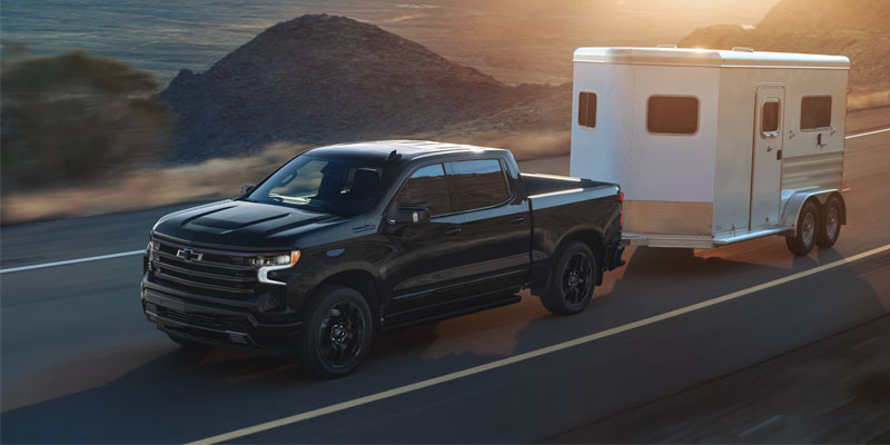 Black Chevrolet Silverado 1500 towing a white horse trailer on a scenic road during sunset.