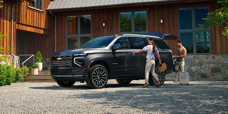 A woman opens the door of a Chevrolet SUV while a man prepares luggage outside a cabin.