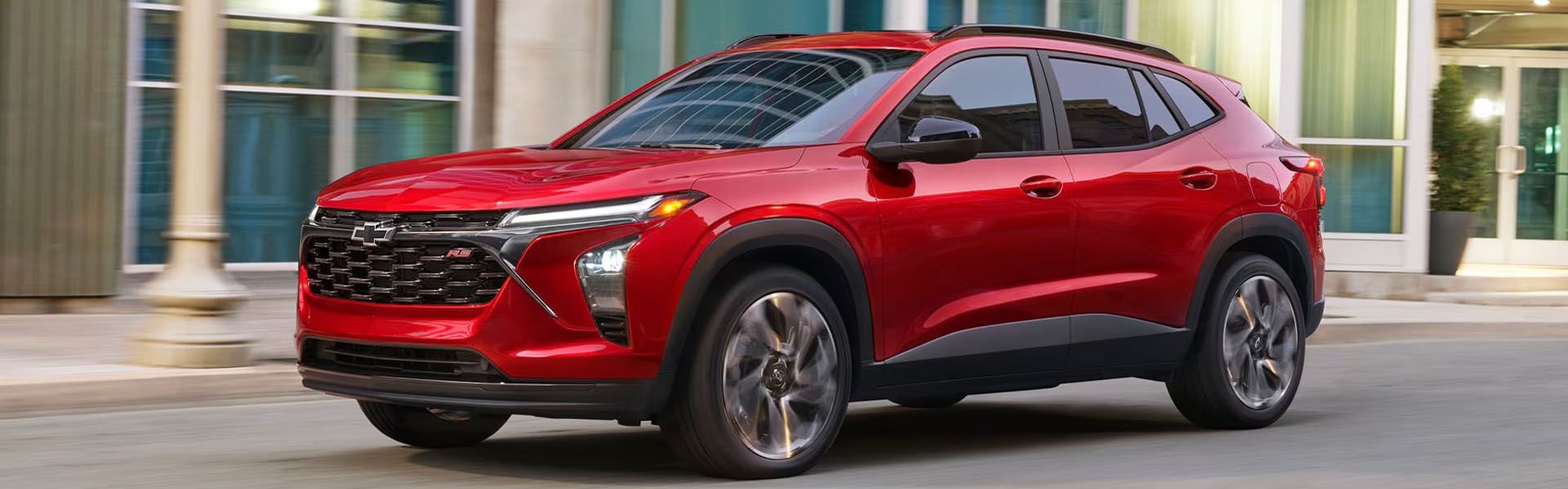 2026 Chevrolet Trax in red parked in a city setting with modern buildings in the background.